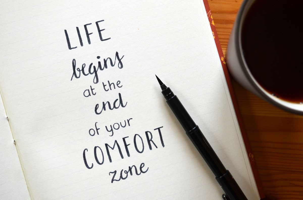 LIFE BEGINS AT THE END OF YOUR COMFORT ZONE written in notebook on wooden desk with cup of coffee and pen LIFE BEGINS AT THE END OF YOUR COMFORT ZONE written in notebook on wooden desk with cup of coffee and pen